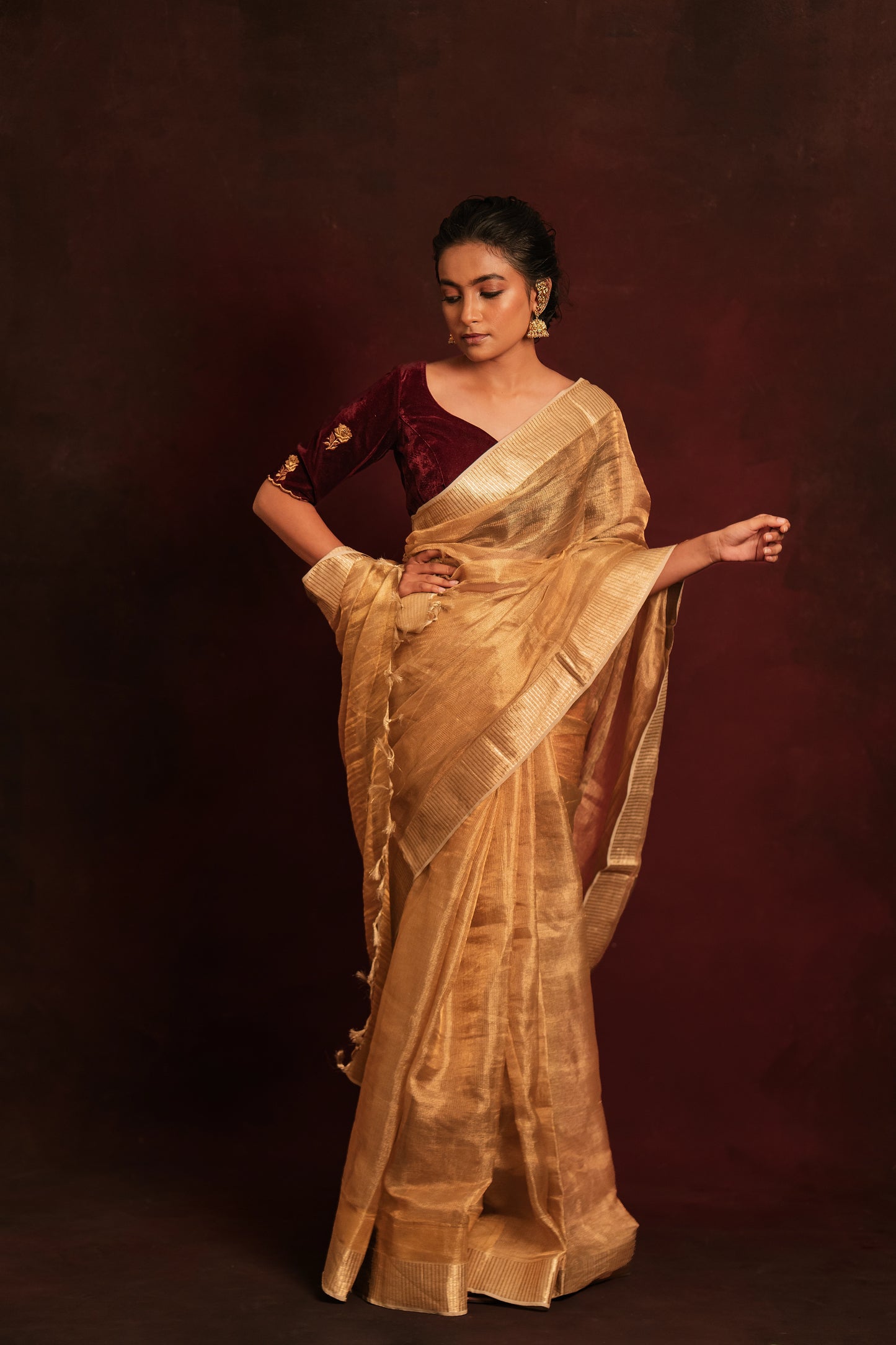 Model Krisha posing for full profile in a tissue saree in gold paired with an embroidered velvet blouse in dark maroon by team AKloset.