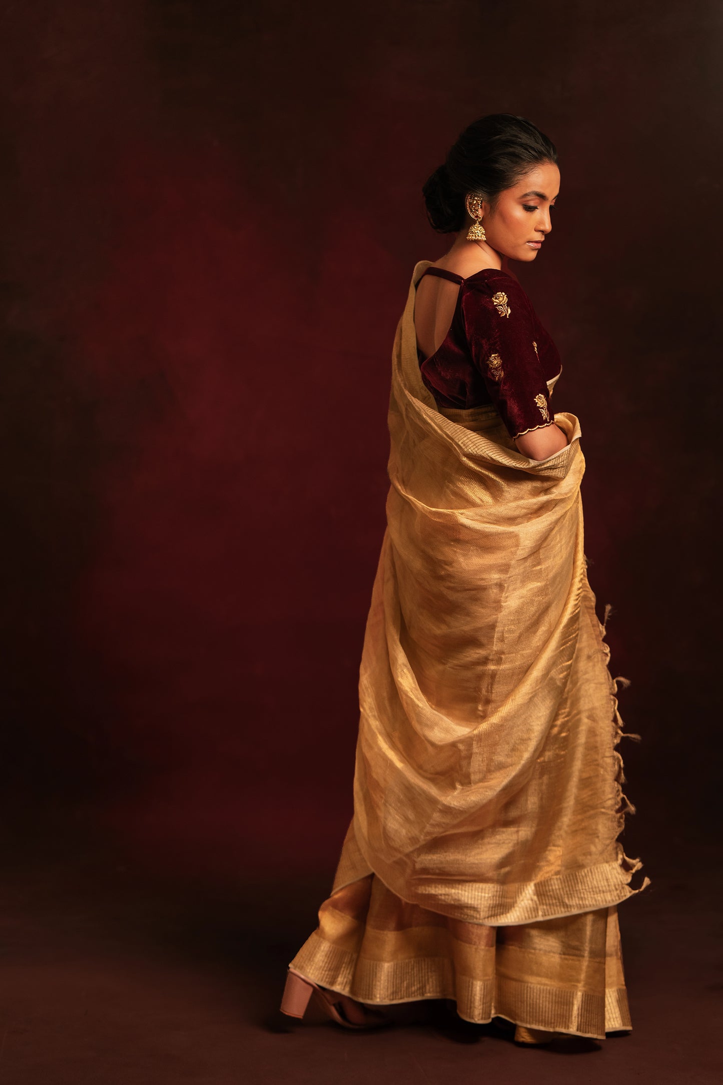 Model Krisha posing for full side profile in a tissue saree in gold paired with an embroidered velvet blouse in dark maroon by team AKloset.