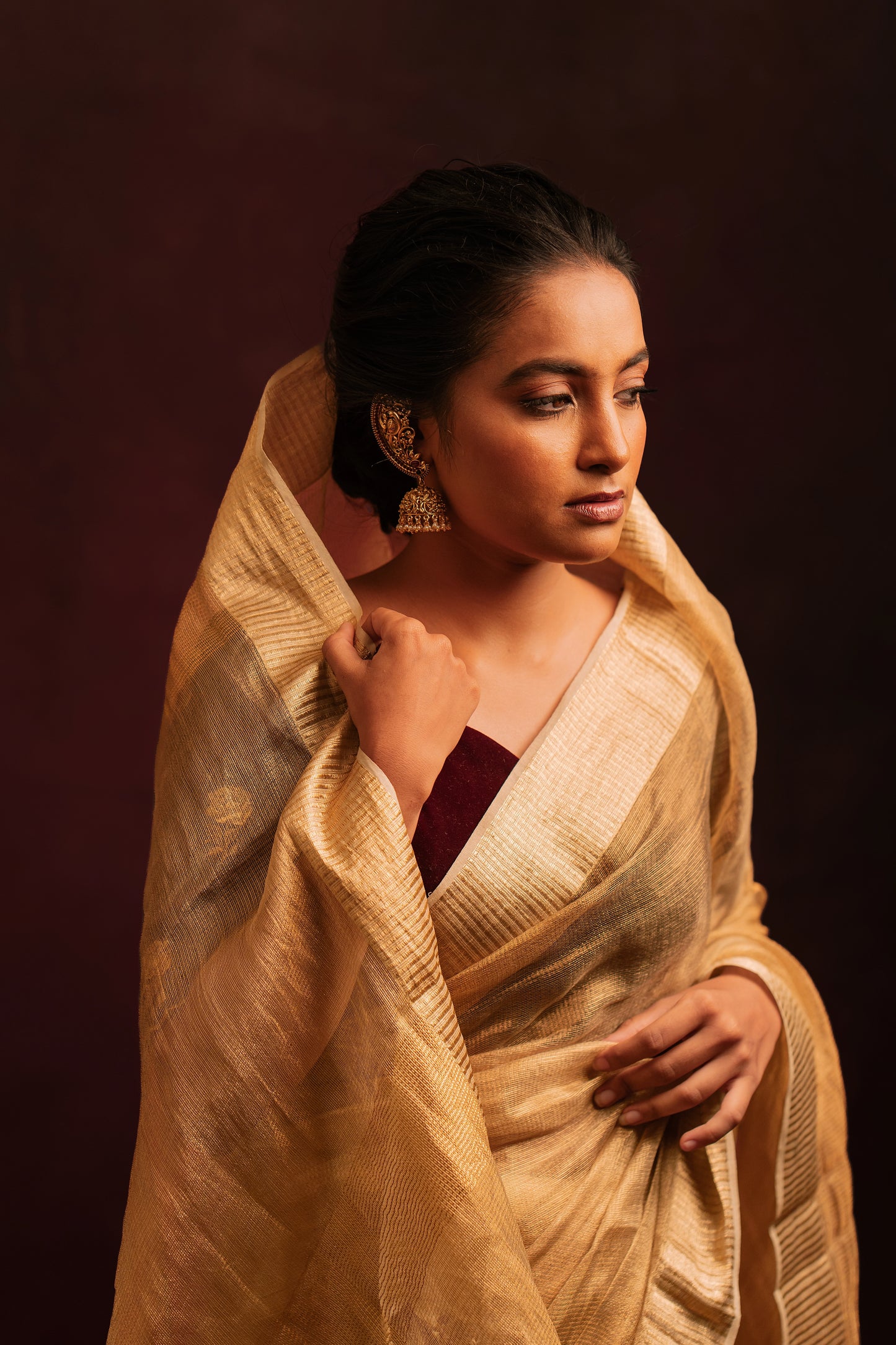 Model Krisha posing for photoshoot in a tissue saree in gold paired with an embroidered velvet blouse in dark maroon by team AKloset.