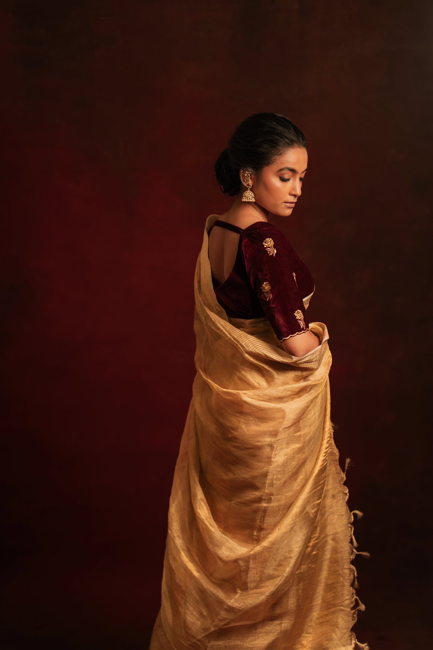 Model Krisha posing for side profile in a tissue saree in gold paired with an embroidered velvet blouse in dark maroon by team AKloset.