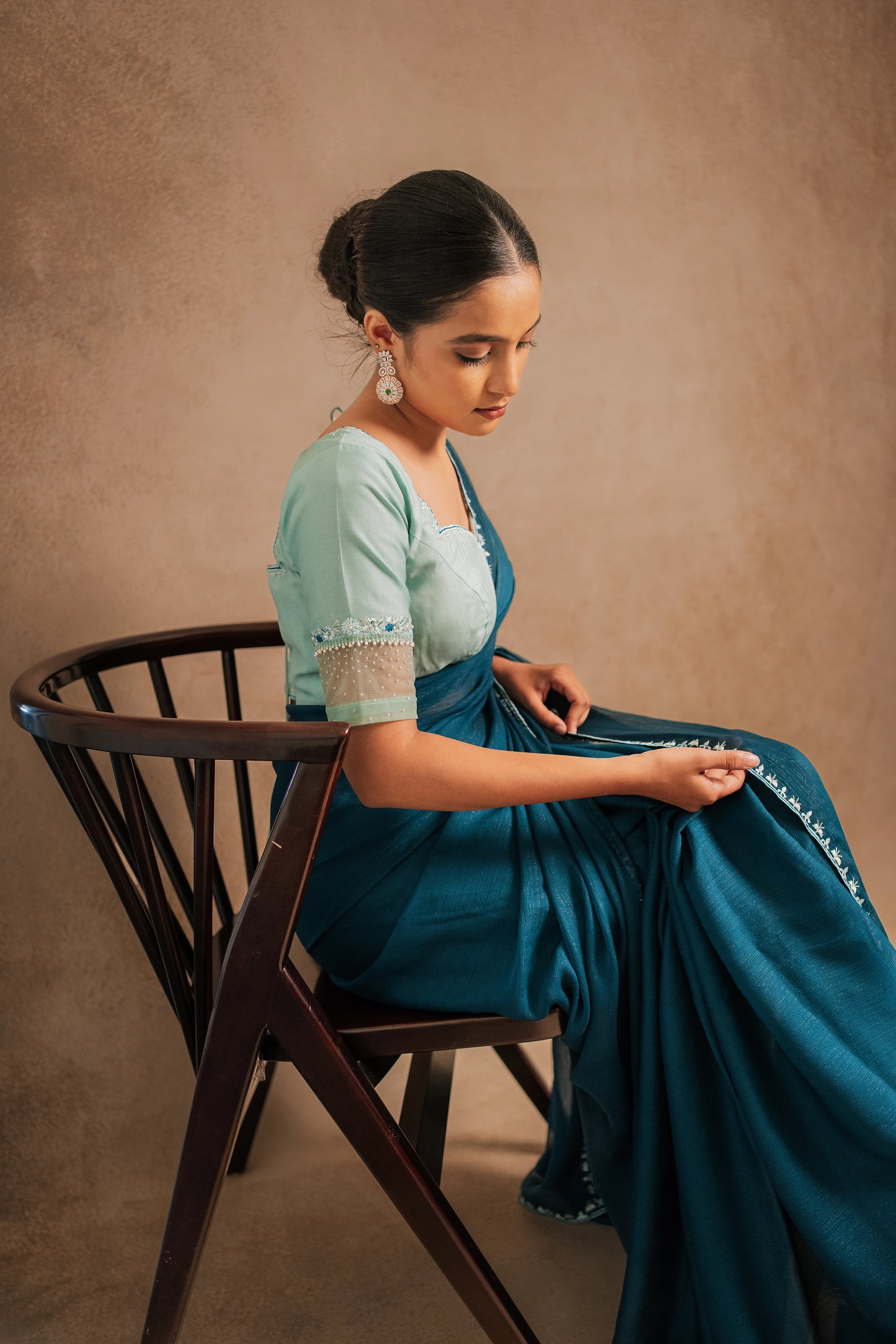 Model Krisha posing seated in embroidered shimmer chiffon saree in teal paired with a chanderi blouse in muted turquoise with pearl beads detailing by team AKloset.